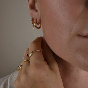 Close-up of a person wearing gold hoop earrings and rings on a neutral background