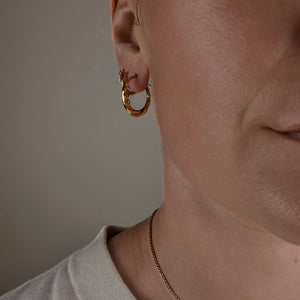 Close-up of a person wearing gold hoop earrings and a delicate necklace against a neutral background.