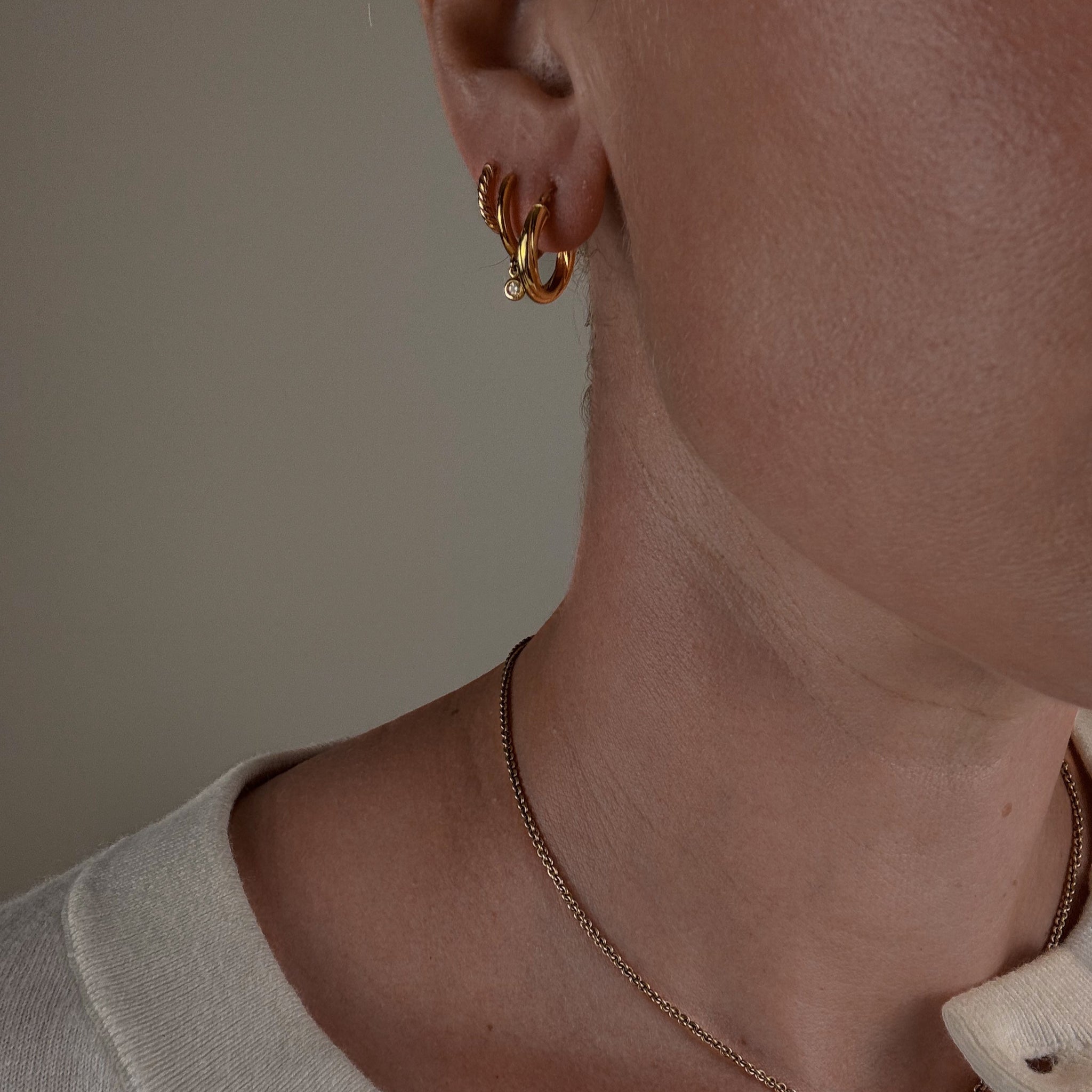 Close-up of a person wearing gold hoop earrings and necklaces against a neutral background