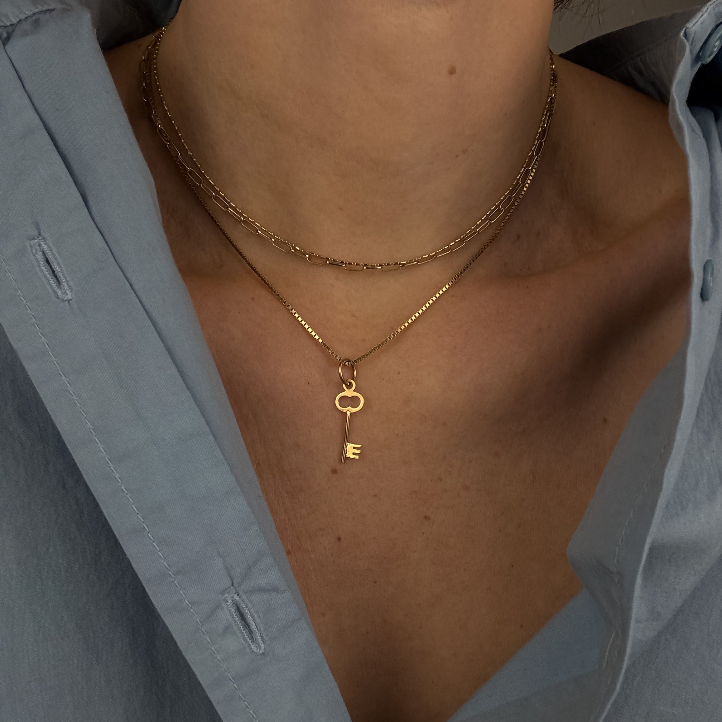 Close-up of a person wearing a gold necklace with a key pendant, partially covered by an open light blue shirt.