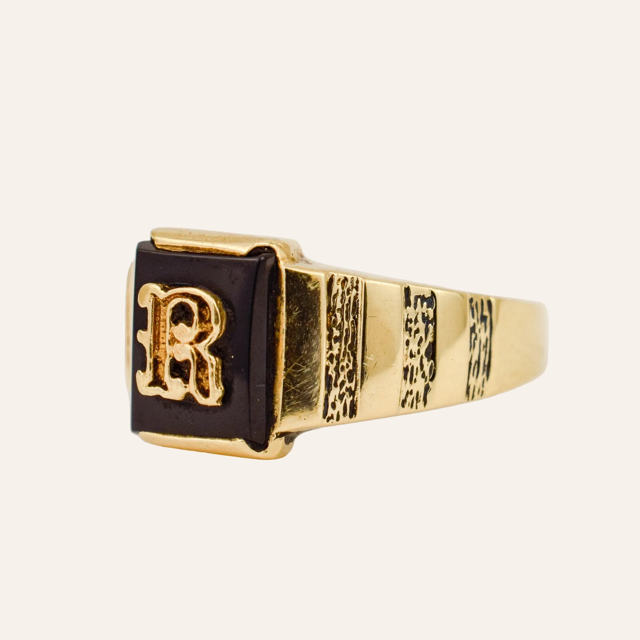 Gold ring with a black square band featuring the letter 'R' on a light beige background